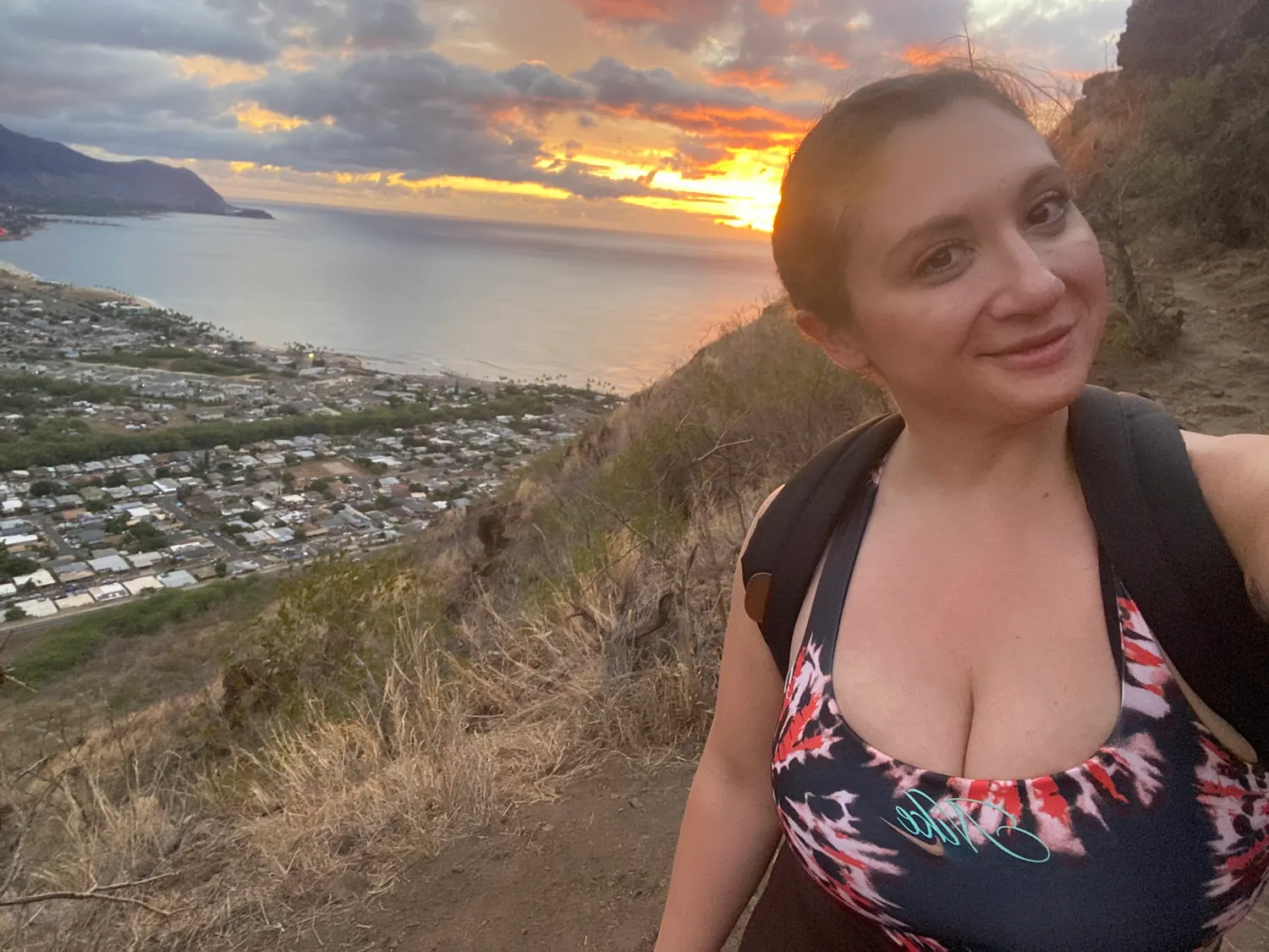 A woman in a printed top taking a picture of herself on a ridge above a town, with the mountain range, the beach and the sea below her and cloud across the sky.