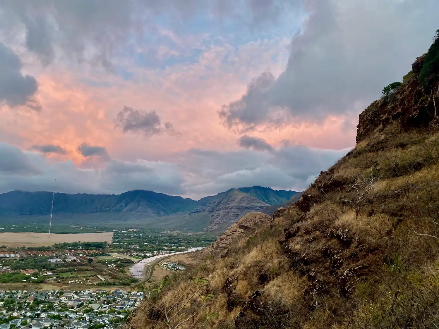 Pink cloud over a valley at dusk, with a town below, a rocky slope at the right and a mountain range across the far side.