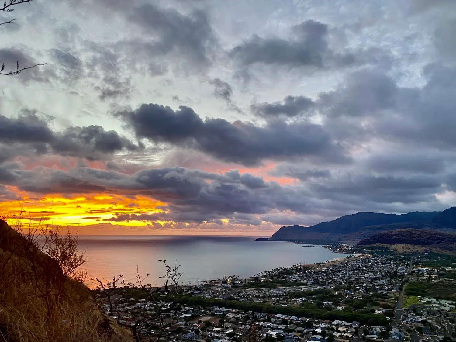 The sun down behind heavy cloud over the sea, with a band of light at the horizon, a dark ridge at the right and the town and coast below.