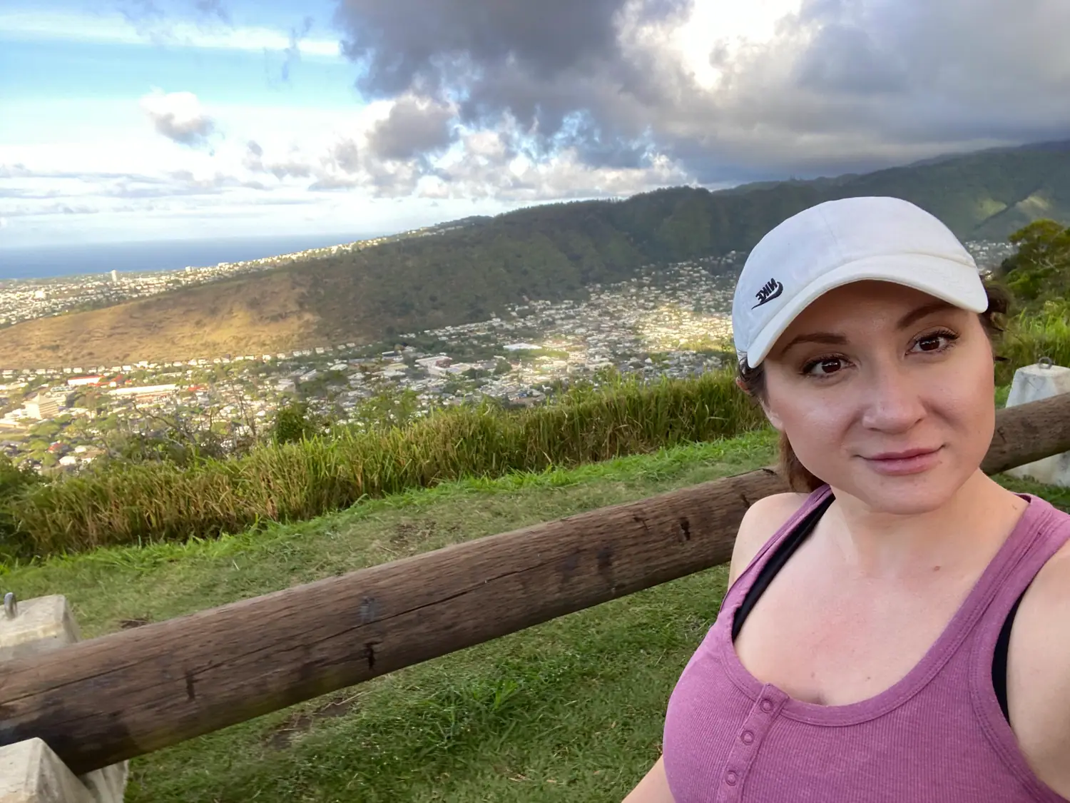 A woman in a white cap and a pink top taking a picture of herself on a green lawn above a city, with a wooden rail across the foreground and the sea and skyline beyond.