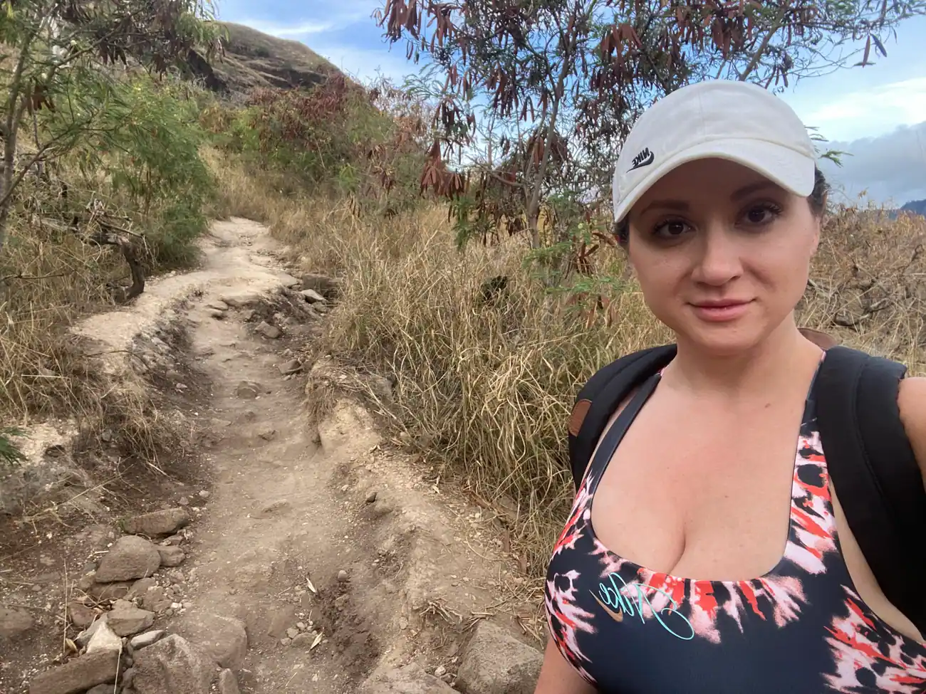 A woman in a white cap and a printed top taking a picture of herself on a rocky dirt trail on a dry hillside, with scrub and a bank of dry grass behind her.