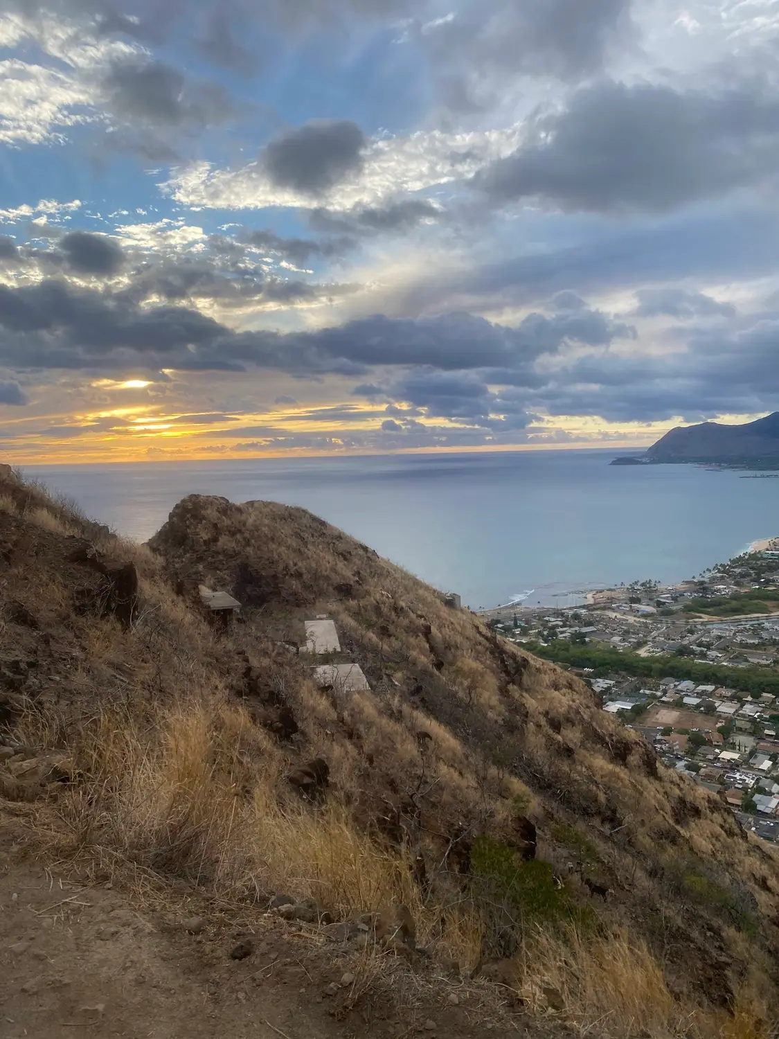 A dry hillside with a low concrete structure on it, seen above a bay, a headland and a town in low sun.