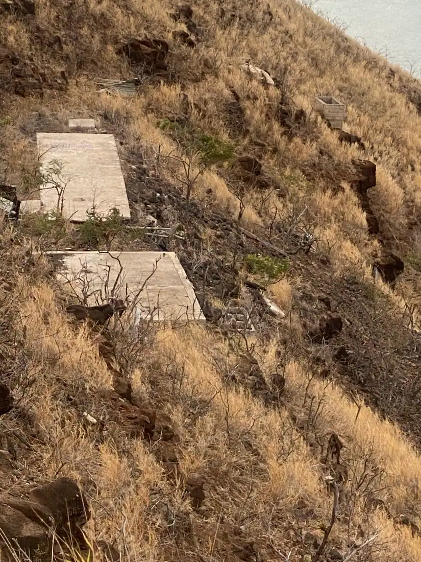 A steep hillside of dry grass and rock with two pale concrete slabs on it and a sliver of the sea at the top right.
