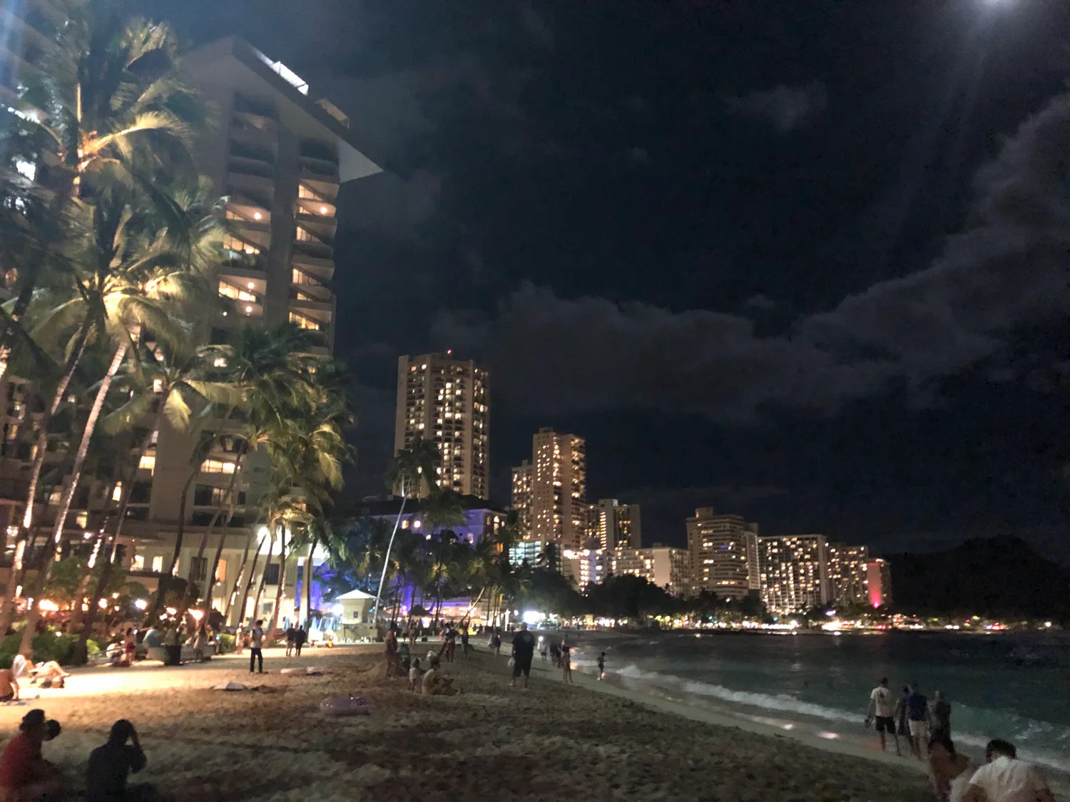 Palm trees on a night beach with lit hotel towers behind, and people walking and sitting along the sand at the water's edge.