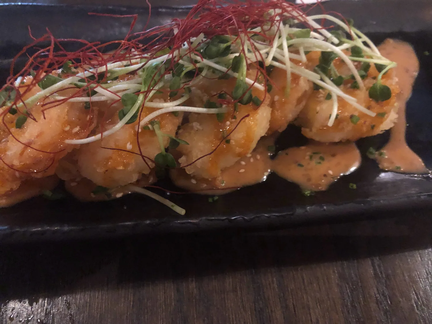 A plated dish of fried shrimp dressed with a pale sauce, shredded greens and fine red strands, on a dark slate plate on a wooden table.