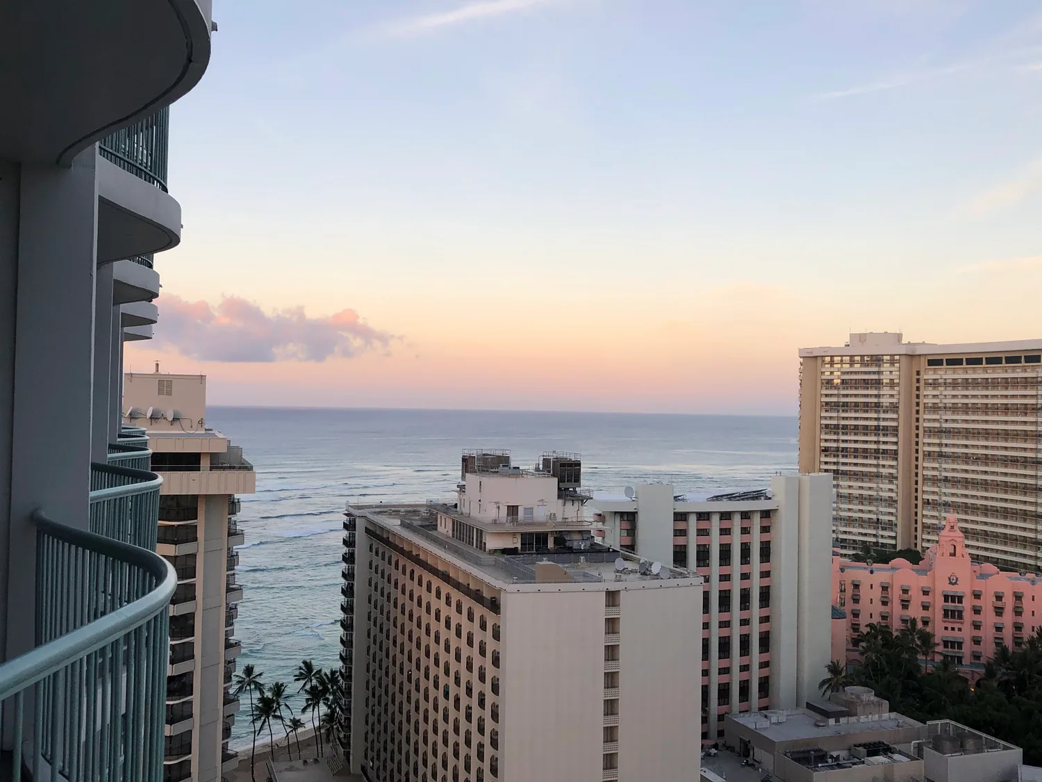 A sea view at first light from a high balcony over hotel roofs, with a curved tower at the left edge, a long striped tower behind and a small pink building below.