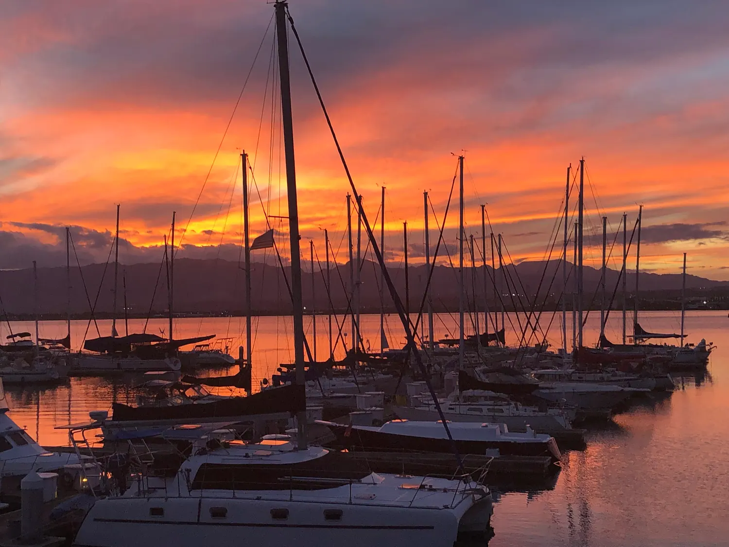 A marina of moored yachts and masts at sunset, the sky burning orange above a dark ridge of hills, with boats in the foreground.