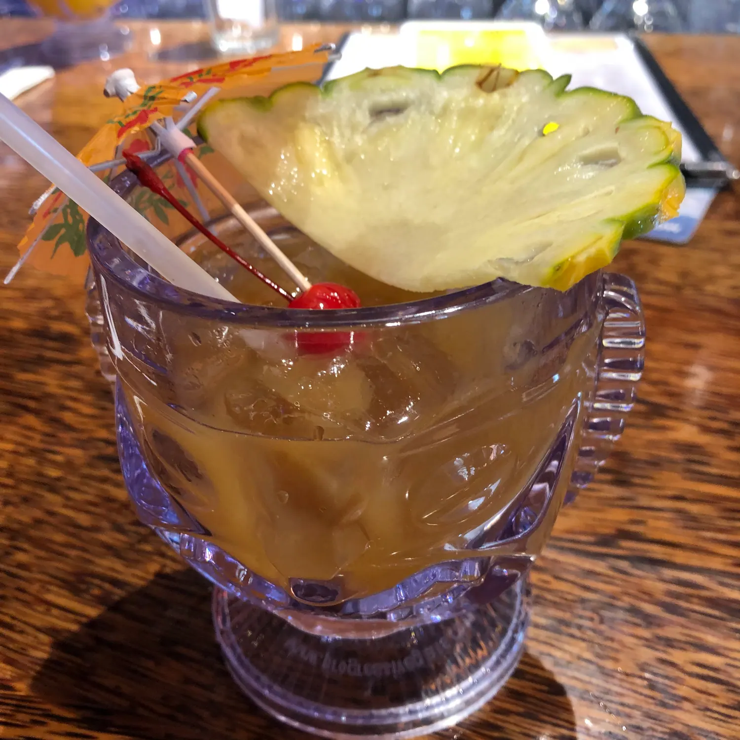 A tall glass of iced drink on a wooden table with a pineapple wedge, a cherry and two straws, and a blurred plate and menu above it.