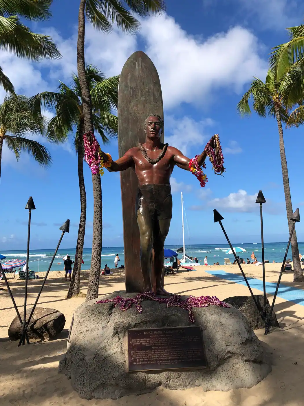 A bronze figure of a man standing with a surfboard upright behind him on a stone boulder carrying a printed plaque, with palms, sand and the sea behind.