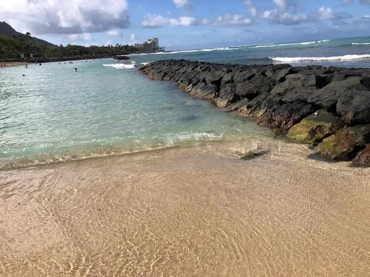 A curving rock breakwater running out into a lagoon along a pale beach, with wet sand in the foreground, swimmers in the water and a headland and buildings along the shore beyond.