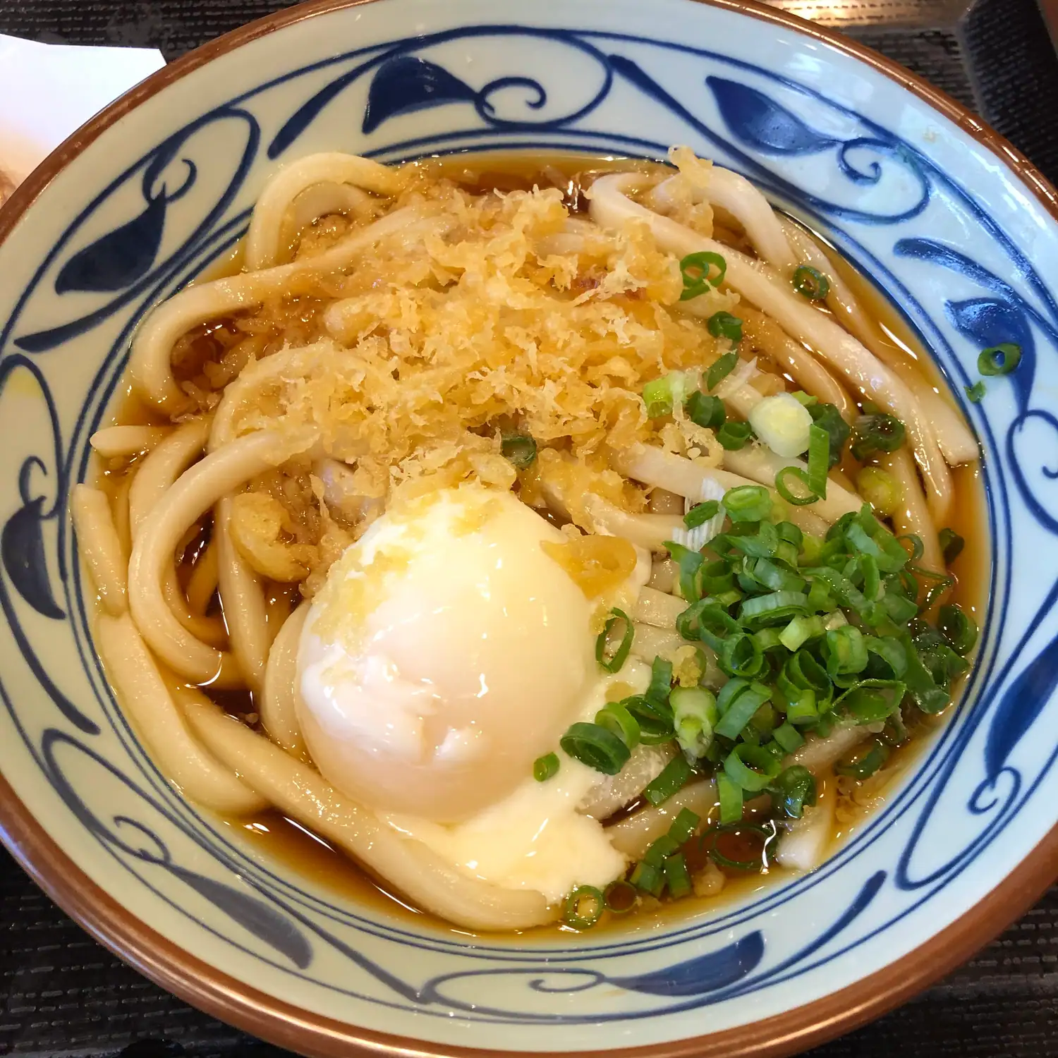 A bowl of thick noodles in broth with a soft egg, chopped greens and crisp flakes, in a blue and white patterned bowl on a dark table.