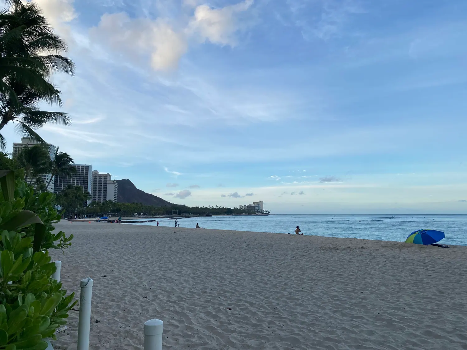 A wide morning beach with buildings and a headland along the shore, a few small figures walking and a folded kayak on the sand.