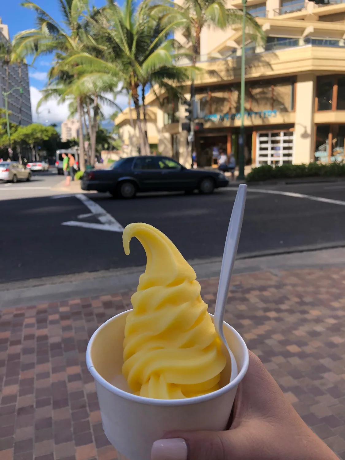 A hand holding a cup of swirled soft serve on a street corner, with a car and a building frontage out of focus behind it.