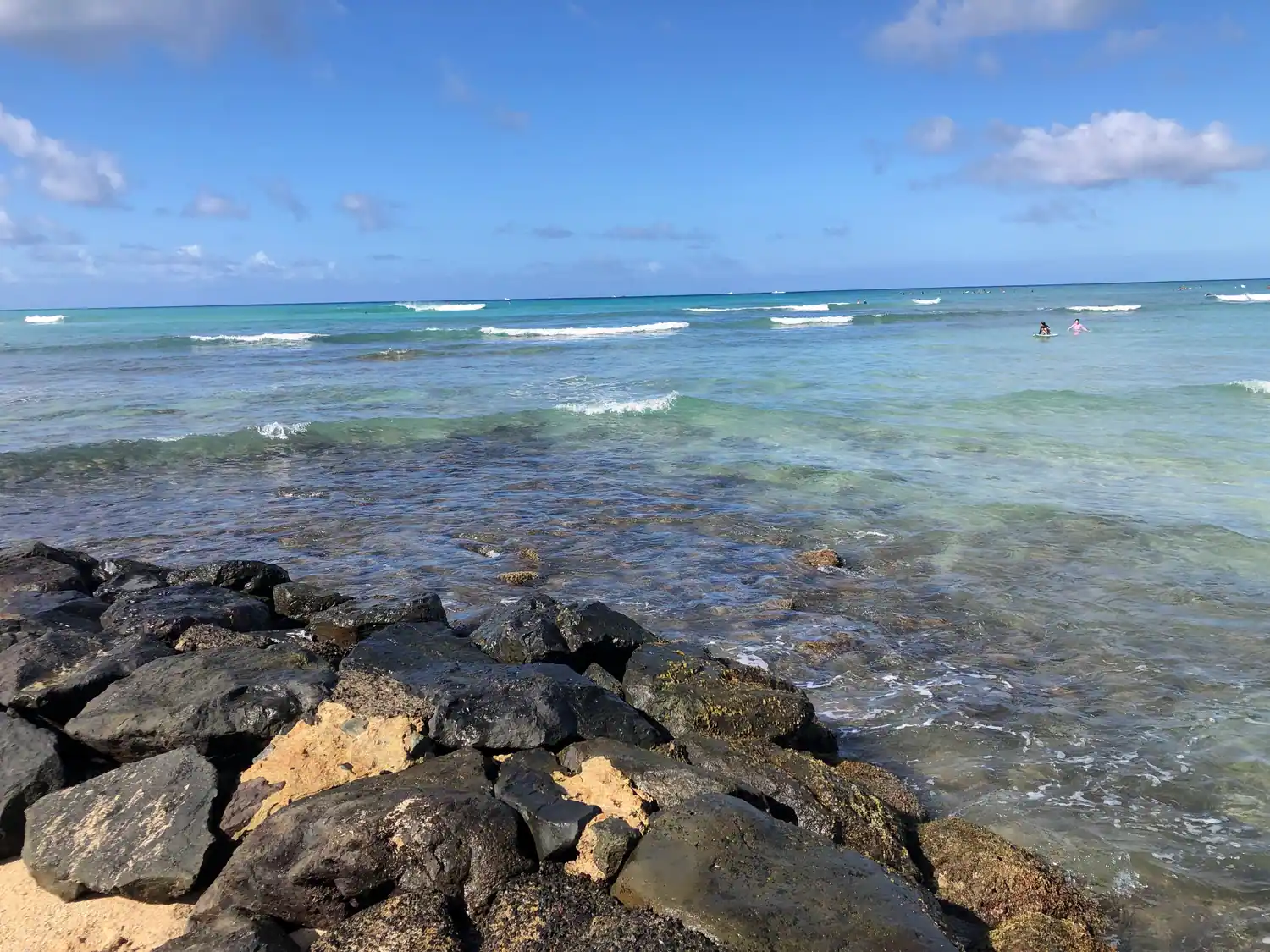 Waves breaking over a dark rock seawall into clear green shallows, with open water beyond.