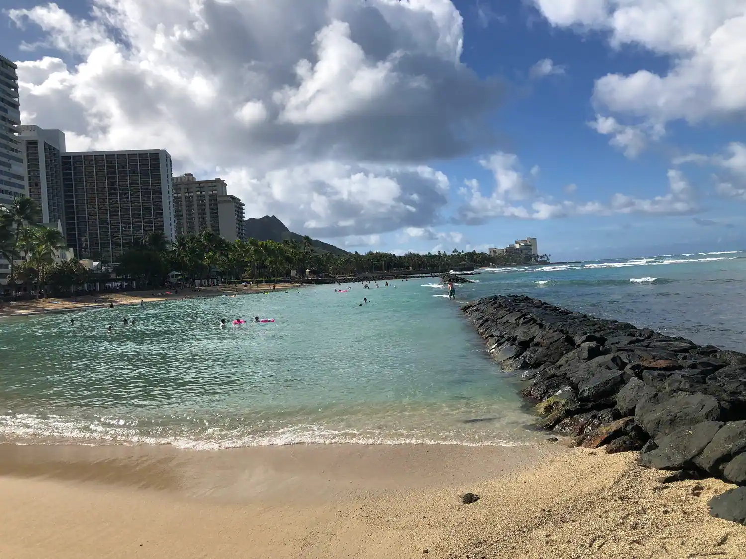 A rock breakwater running out into a lagoon, with swimmers and floats in the water and tall buildings and a headland along the shore.
