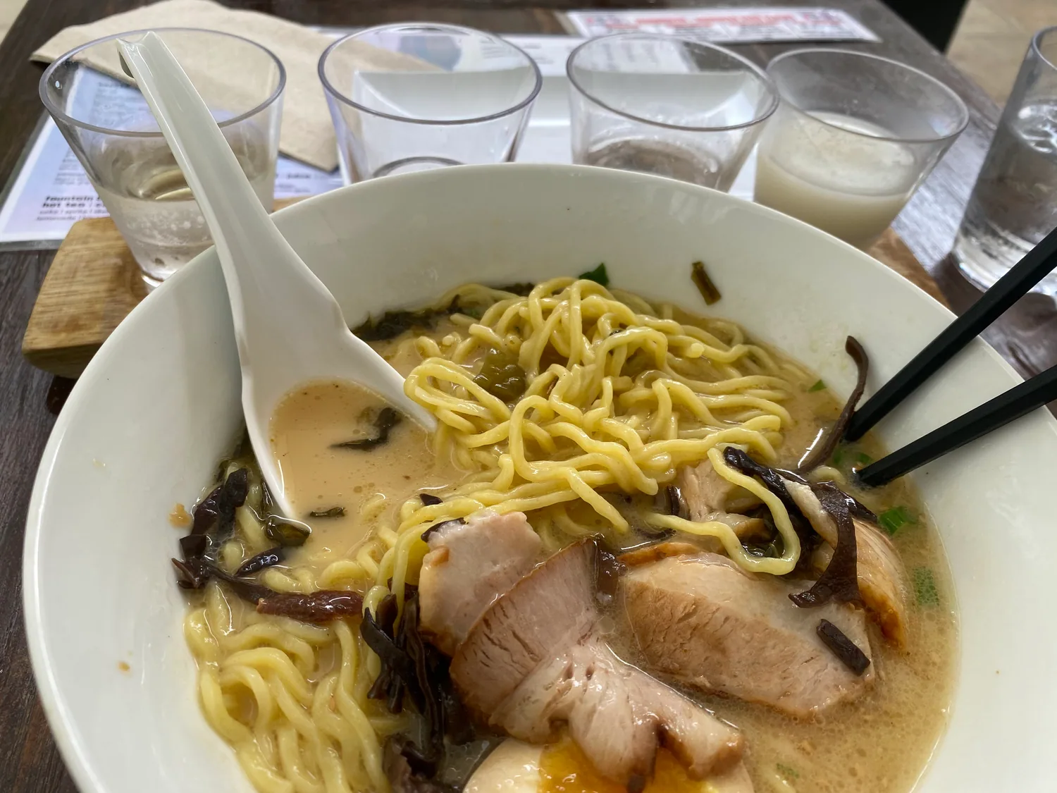 A bowl of noodles with sliced pork and mushrooms in a pale broth on a table, with glasses of water and a menu card beside it.