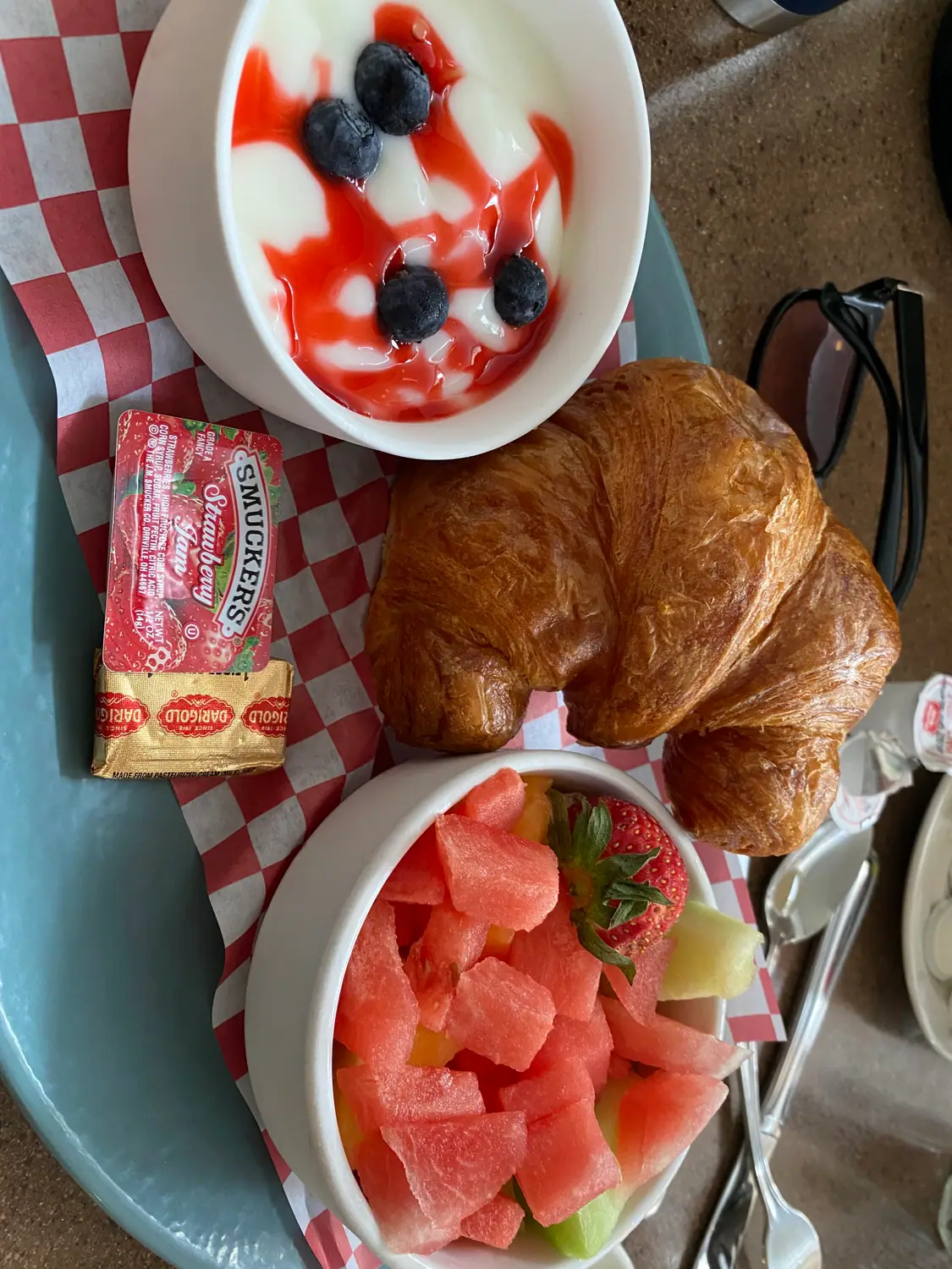 A breakfast tray on a checked cloth with a bowl of yogurt and berries, a croissant, a bowl of melon and a small packet of jam, with sunglasses laid beside it.