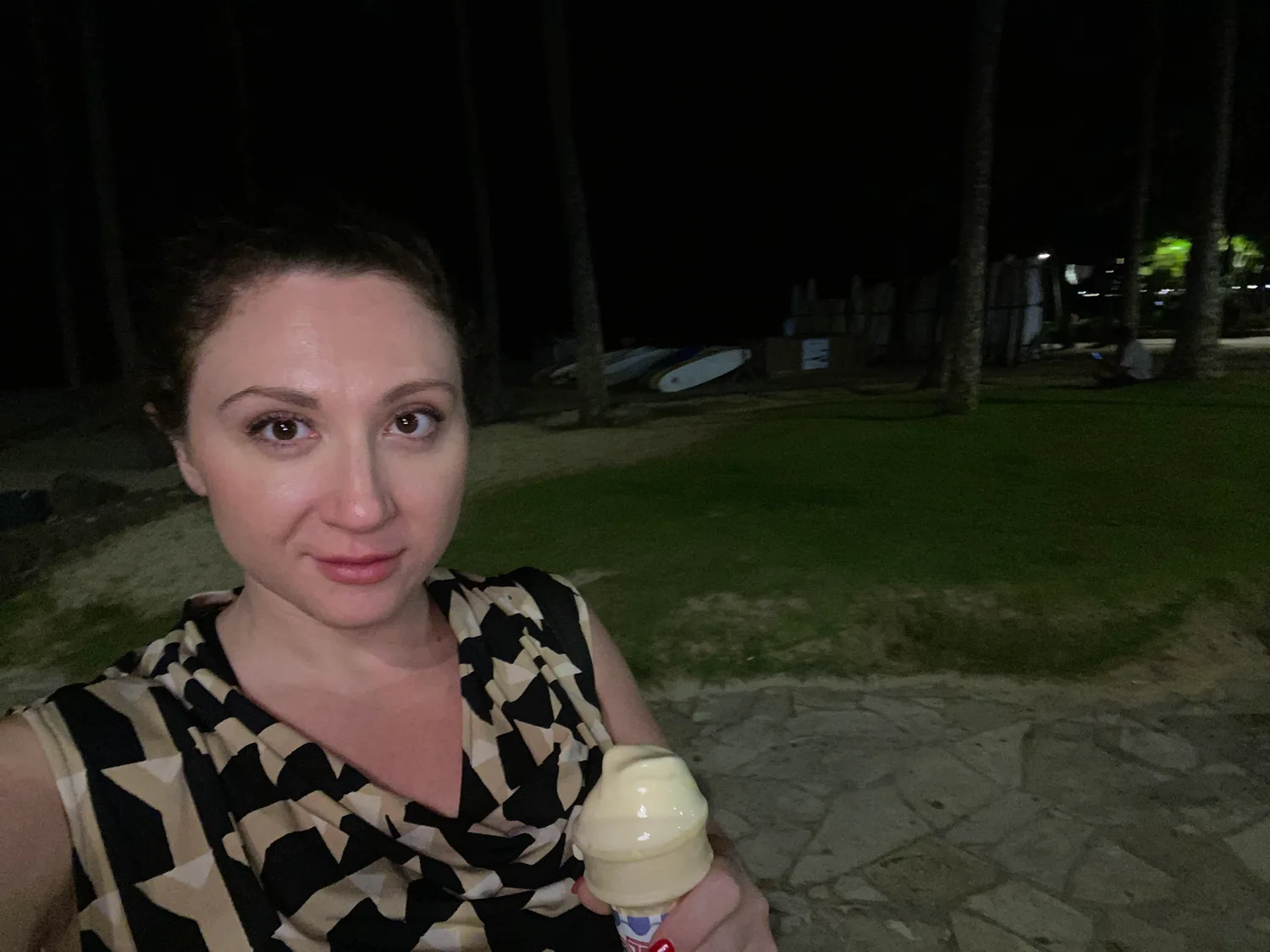 A woman taking a picture of herself at night on a flagstone path with grass, trees and a lamp behind her, holding a soft serve cone up in one hand.