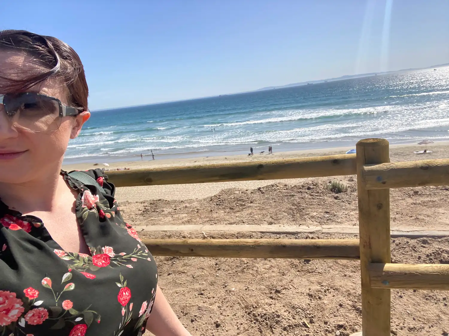 A woman in a dark floral top at a wooden railing above a beach, the ocean and sand behind her.