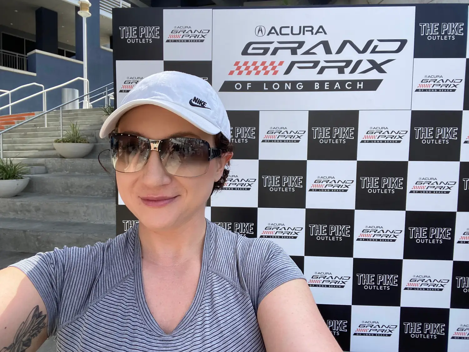 A woman in a white cap and dark sunglasses taking a selfie in front of a wall of repeated printed panels.