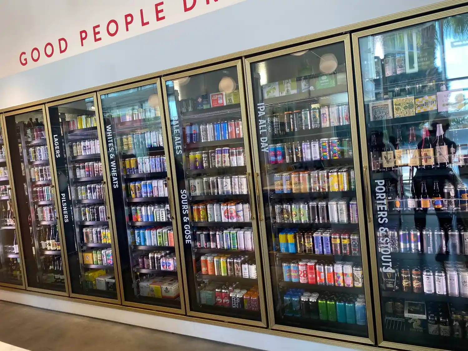 A shop wall of glass fronted cabinets filled with rows of drink cans, painted lettering on the wall above and a bare floor below.