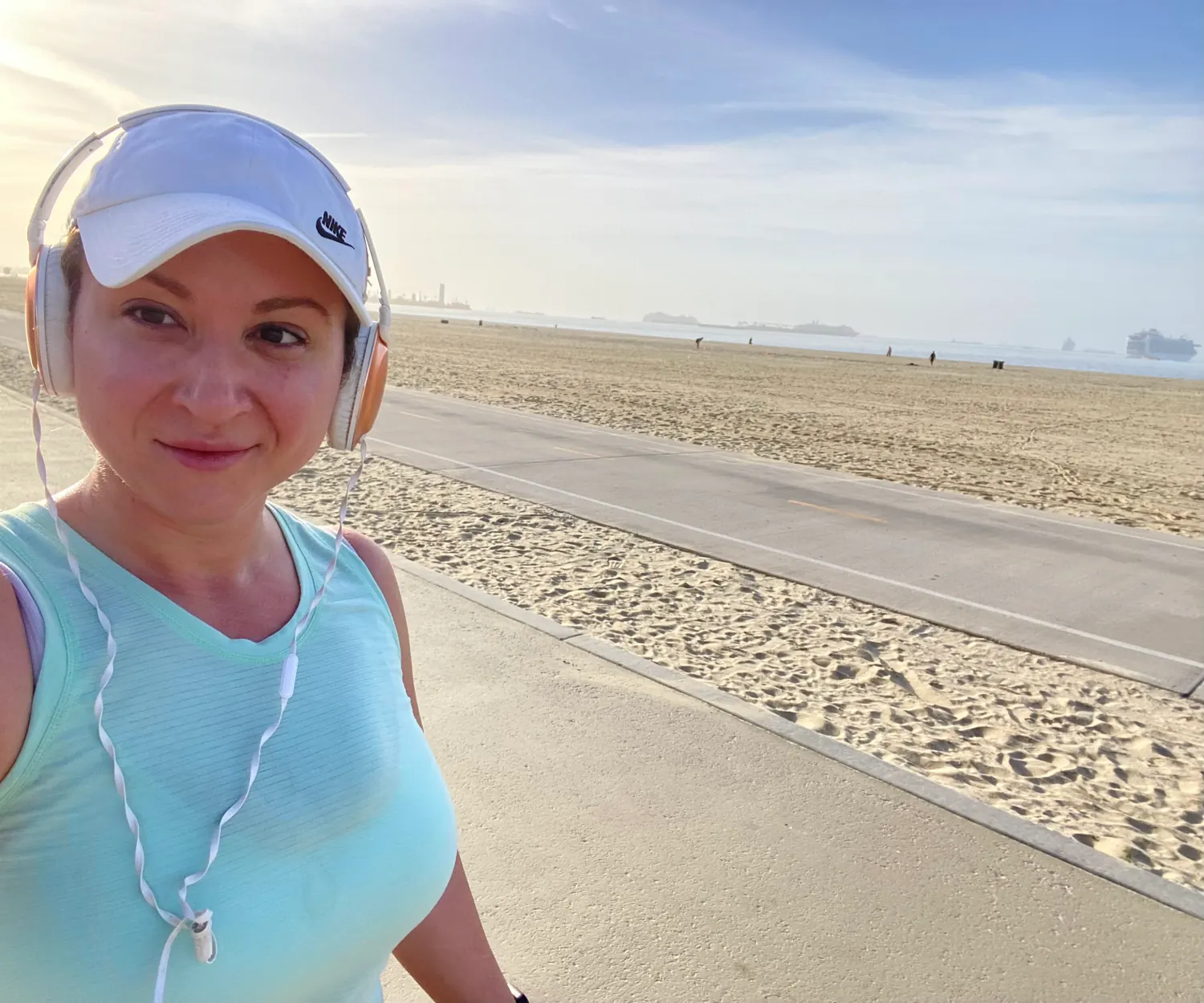 A woman in a white cap and headphones on a path beside the sand, the sea behind her.
