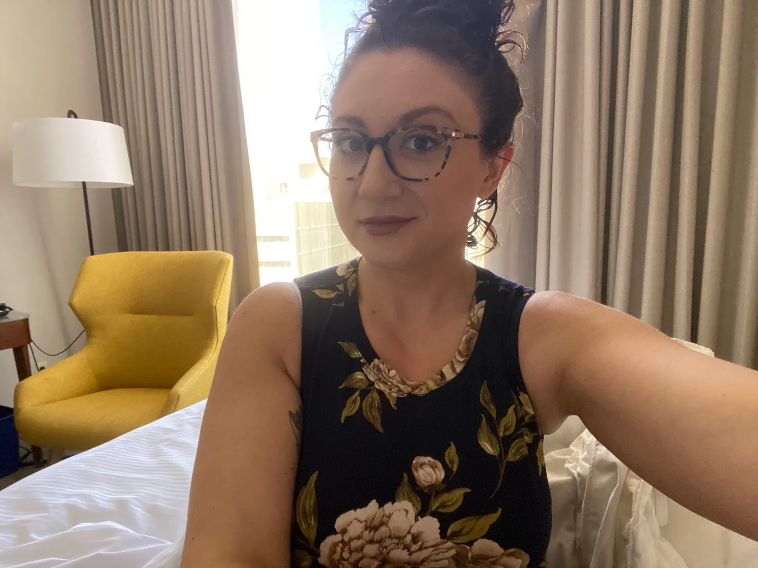 A woman in glasses taking a picture indoors, a bed and a yellow armchair behind her.