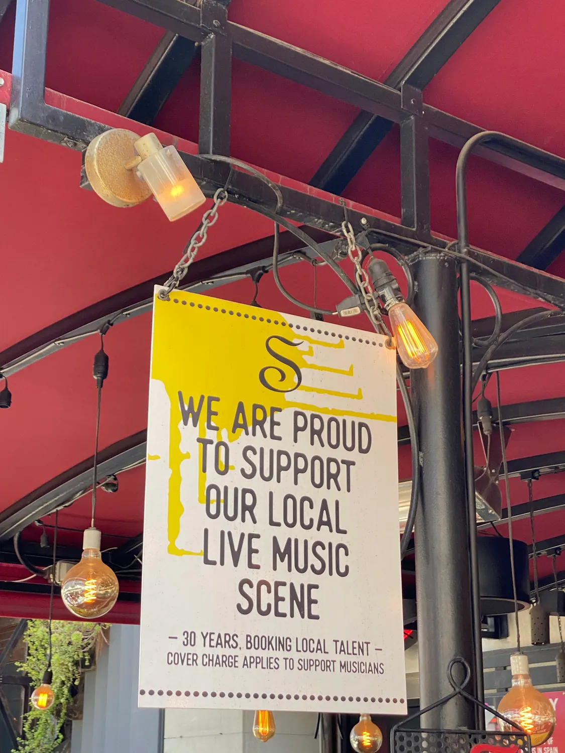 A bright sign with dark lettering hanging from a metal beam beside warm bulbs, red painted steel behind it.