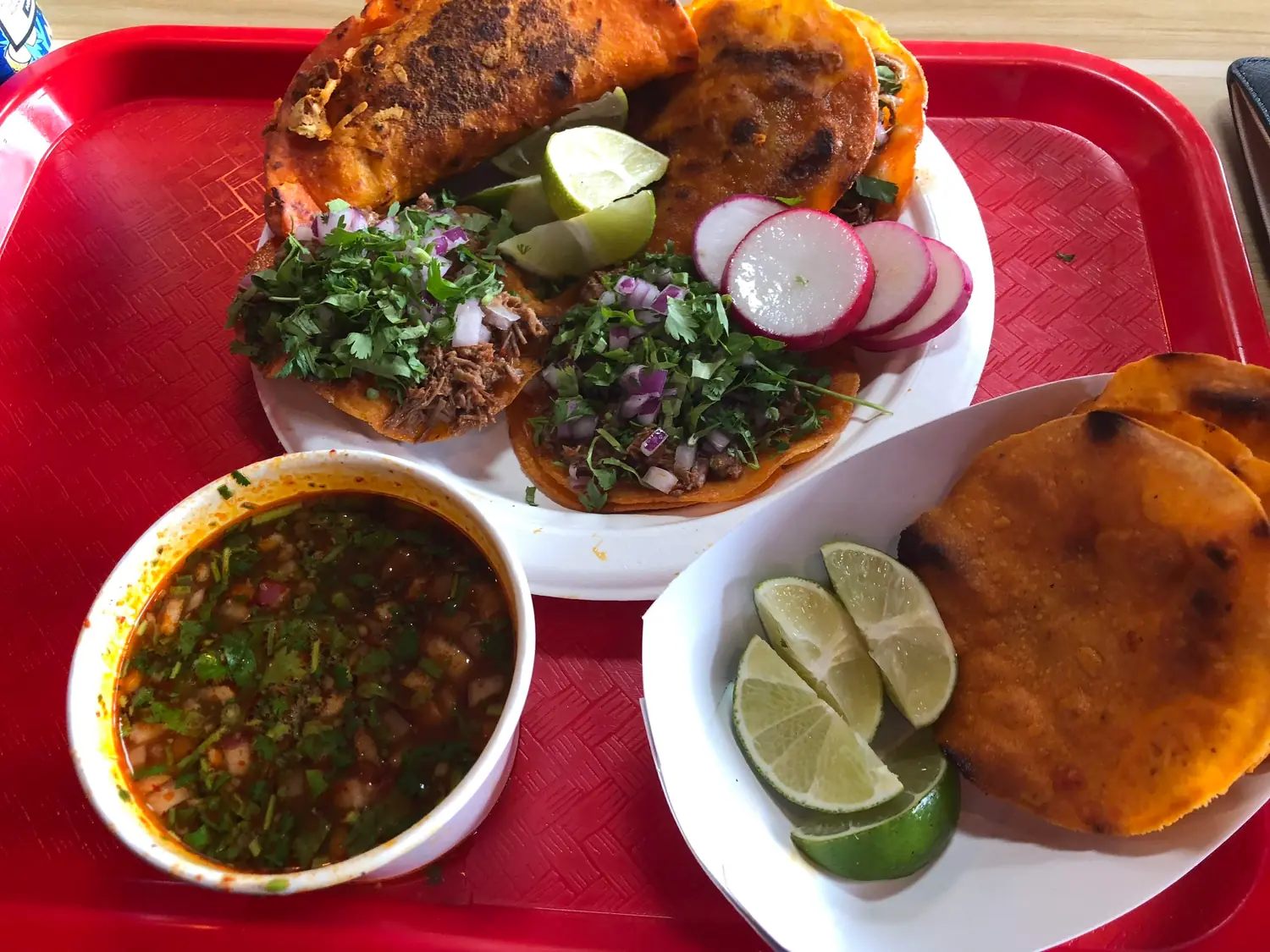 A tray of tacos on two plates with lime wedges and sliced radish, a bowl of green salsa beside them.