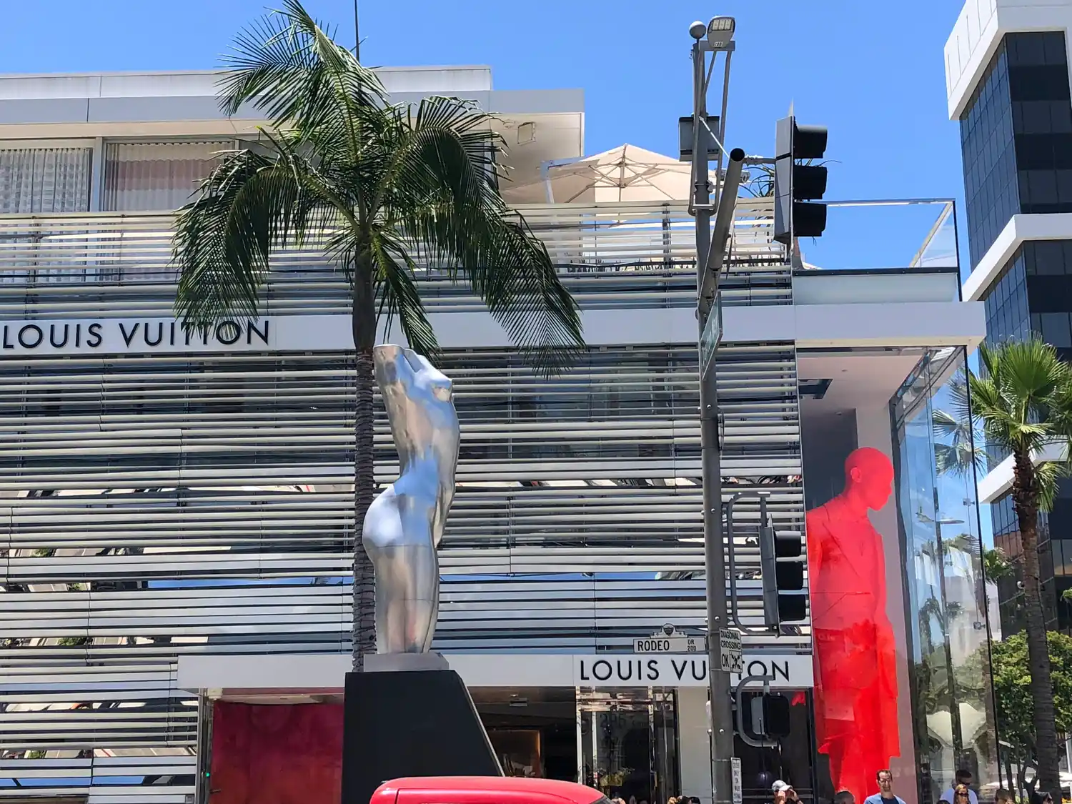 A palm tree and a silver sculpture in front of a pale louvred building on a sunny street corner, traffic lights on the pole beside them.