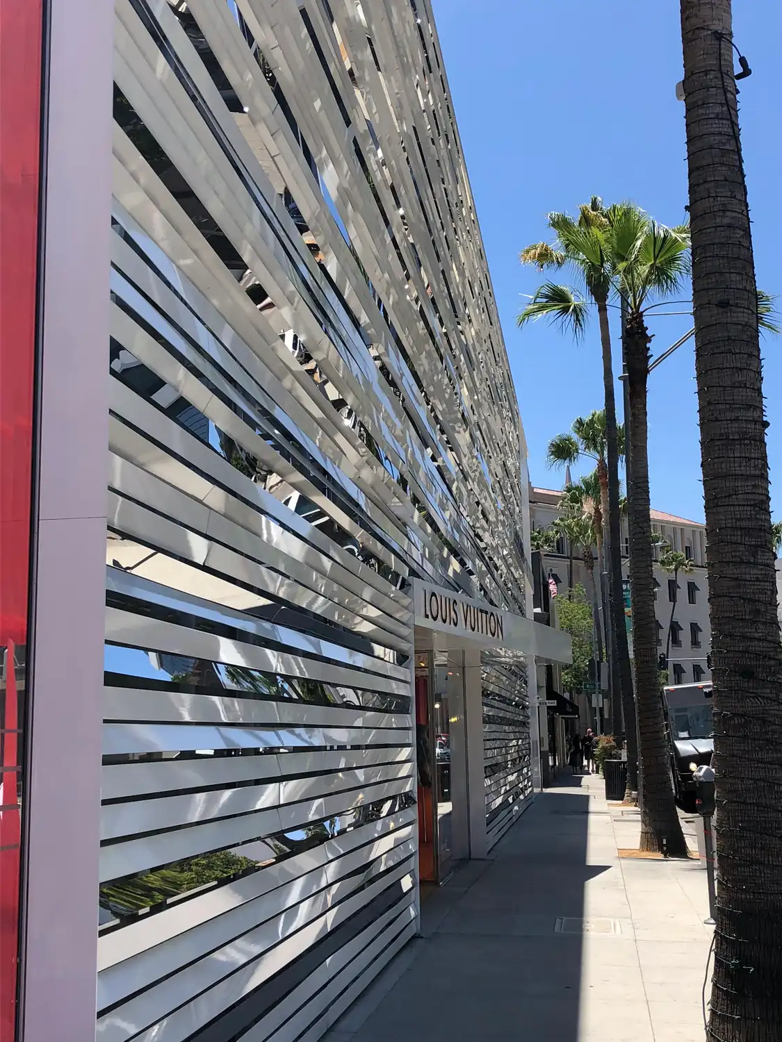 A mirrored louvred wall running along a pavement, palm trunks and a blue sky above, two small figures further down the street.