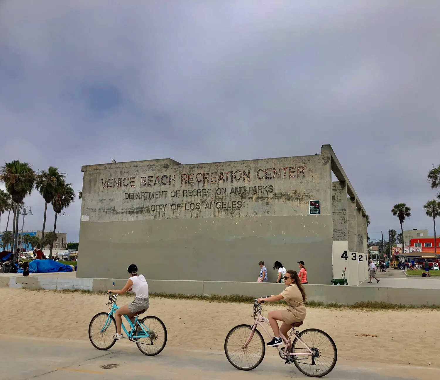 A pale concrete building beside a wide beach path, with two people riding bicycles past it.
