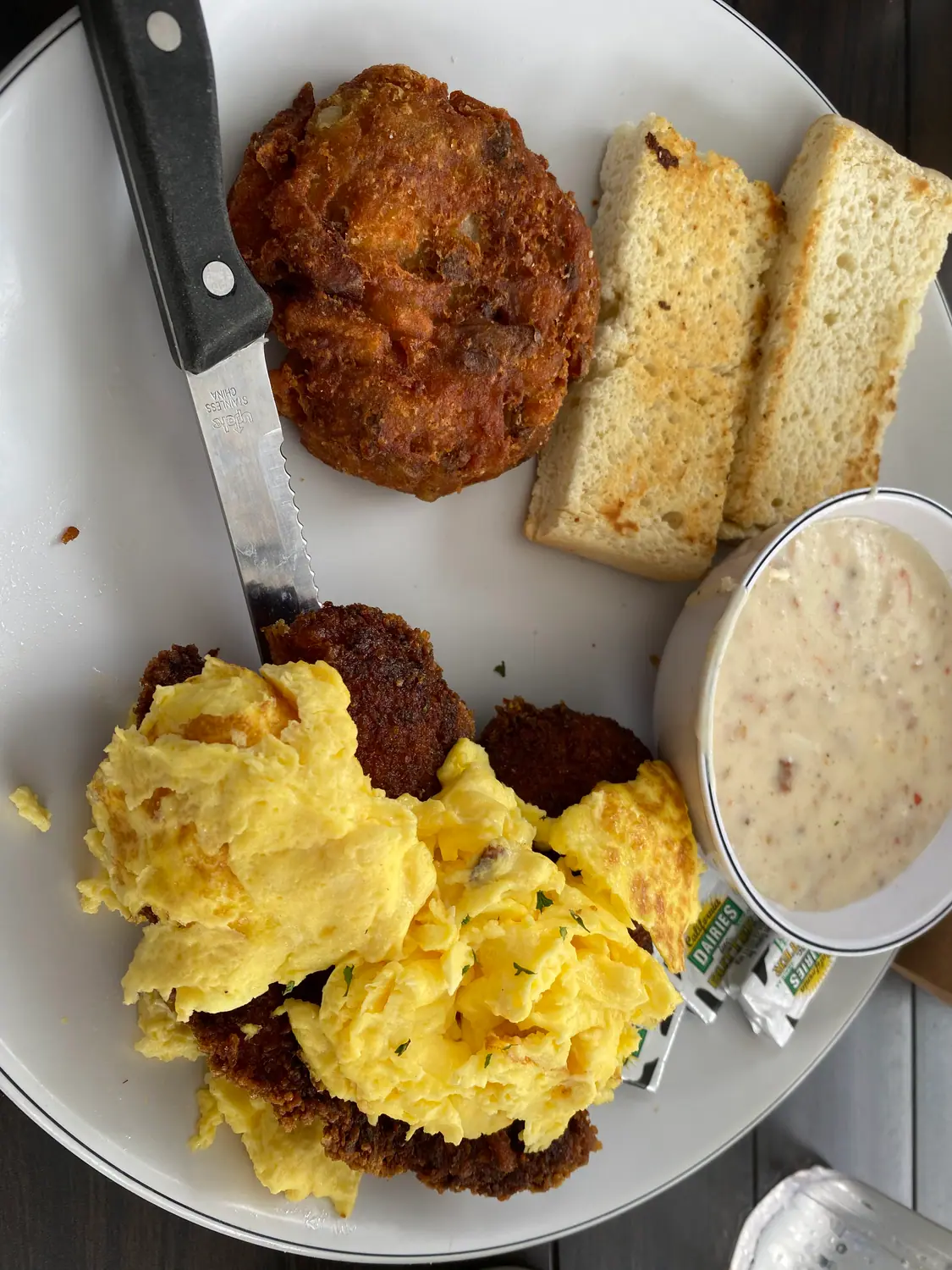 A plate of scrambled eggs, fried potatoes and a biscuit, with a small bowl beside it.