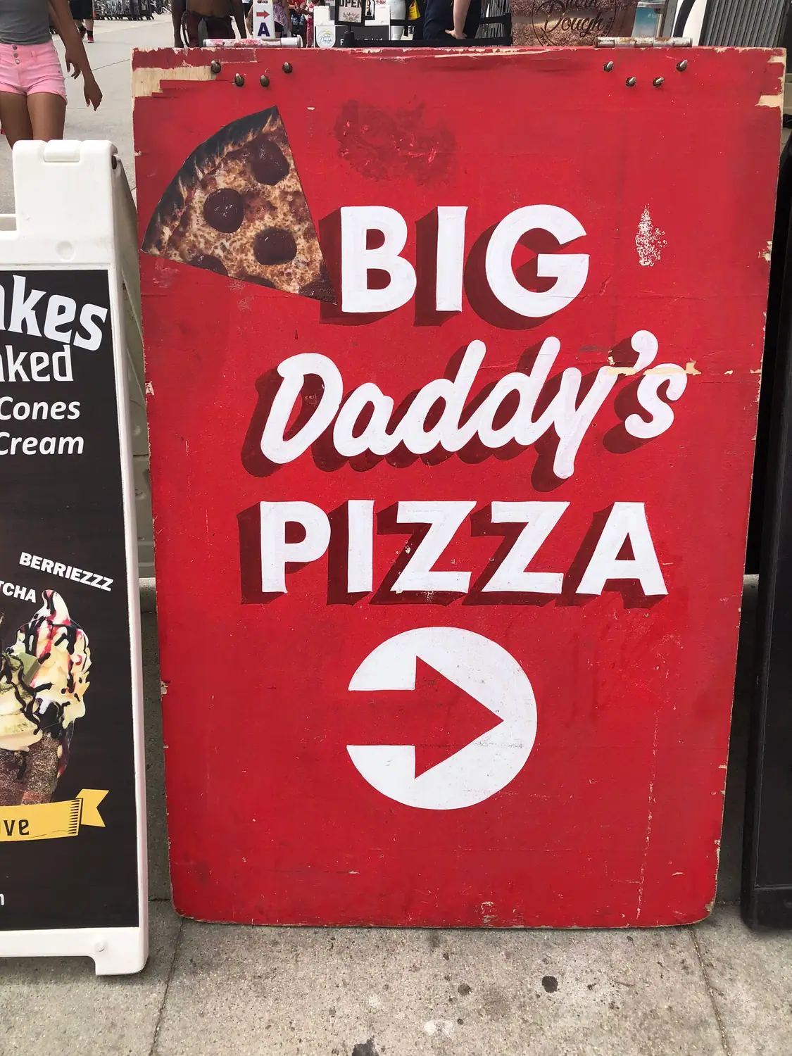 A red painted sign board showing a slice of pizza and a downward arrow, with a smaller sign stand beside it.