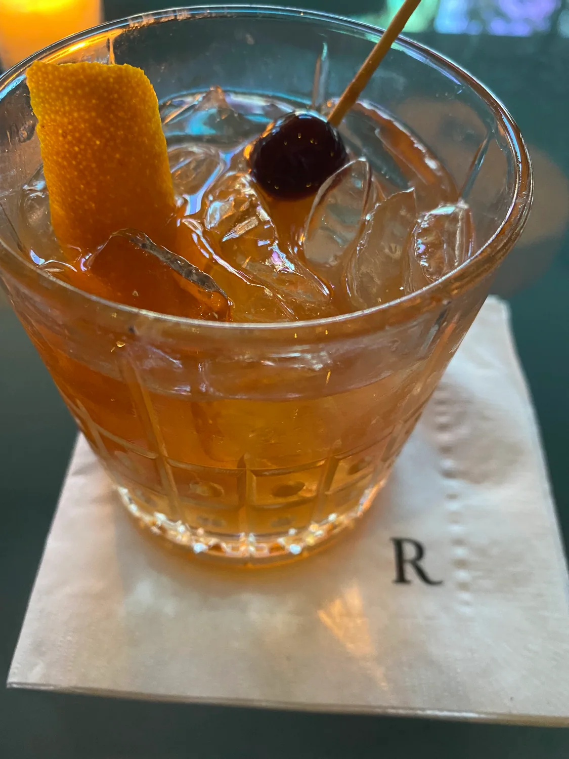 A cocktail in a wide glass with a large ice cube and a twist of orange peel, on a paper napkin.