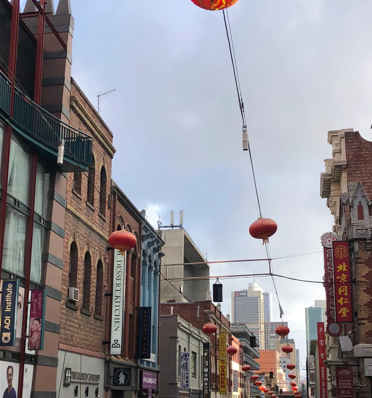 Red lanterns strung on wires above a narrow city lane, brick buildings and shopfronts either side under a grey winter sky.