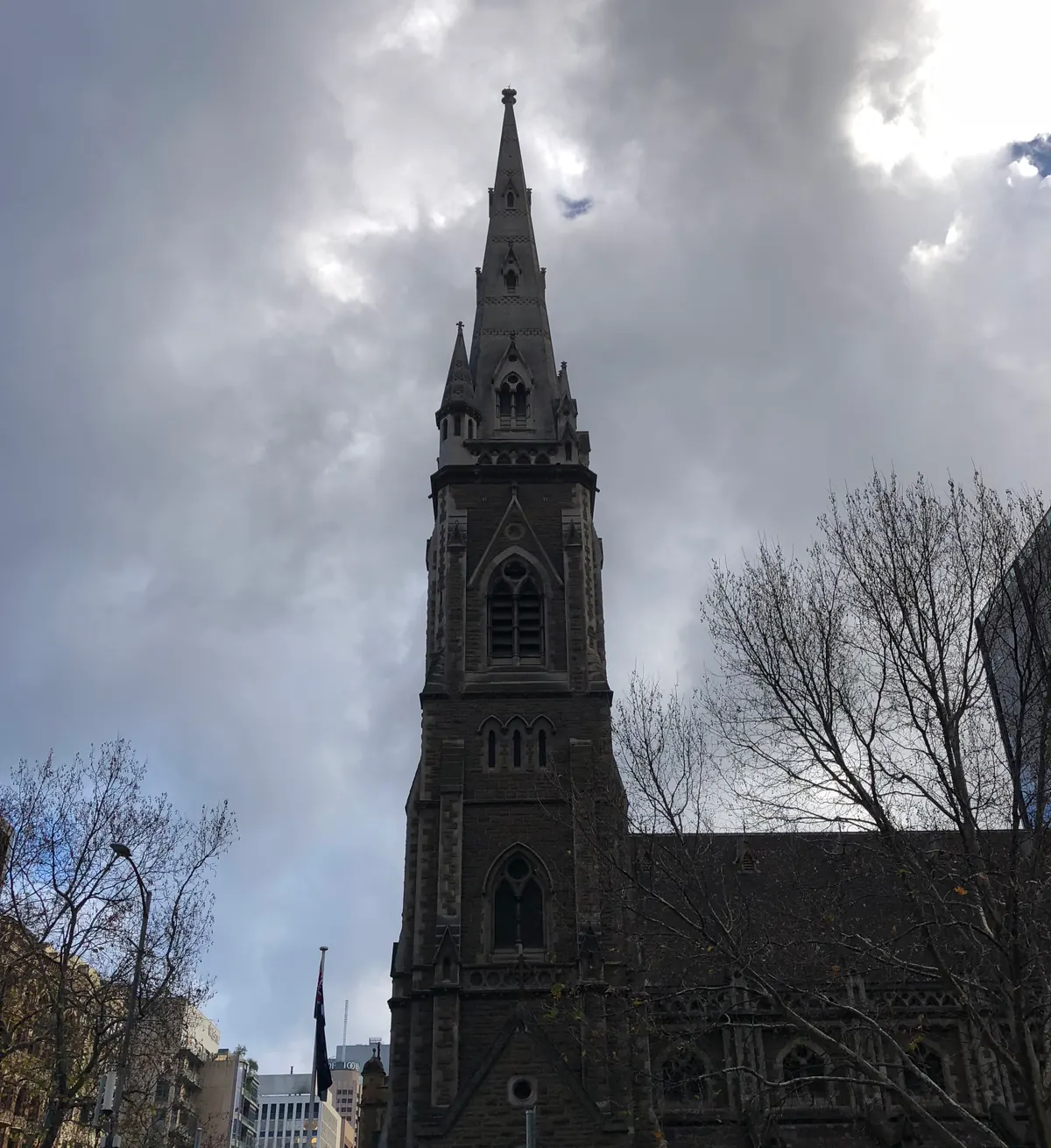 A pale stone spire rising into heavy grey cloud, bare trees around it and the city low behind.