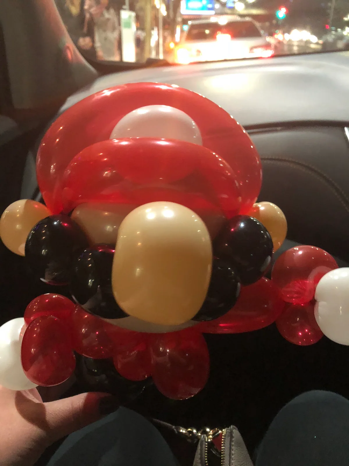 A red, black and cream balloon figure held on a lap inside a car at night, the lit street blurred through the windscreen behind it.