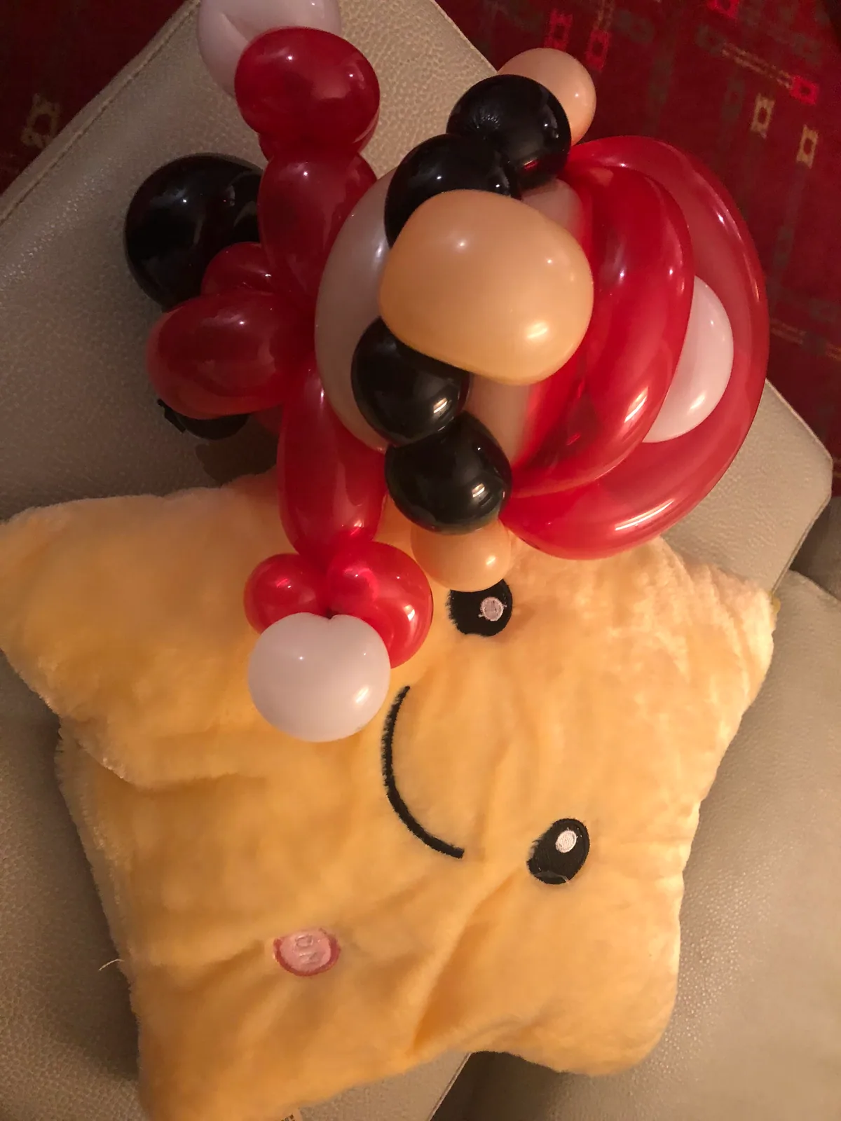 The same balloon figure standing on a soft cushion on a seat indoors, with a patterned carpet behind it.