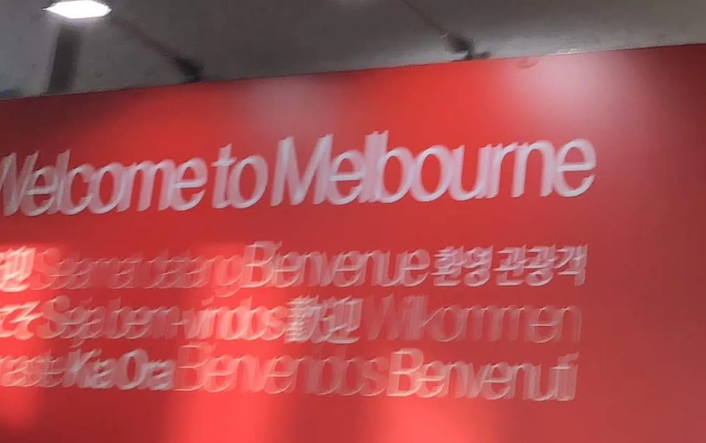 A wide red arrivals panel greeting visitors to Melbourne in a dozen languages, the city's name in large letters across the top and the plain wall above it.