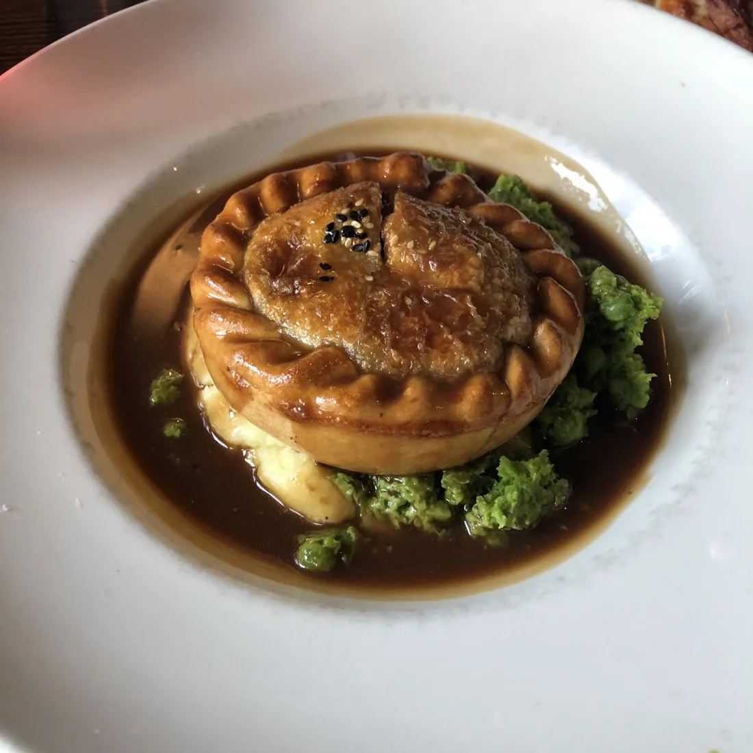 A small round pie with a fluted crust sitting in dark gravy, with mash and mushy peas at the edge of a white plate.