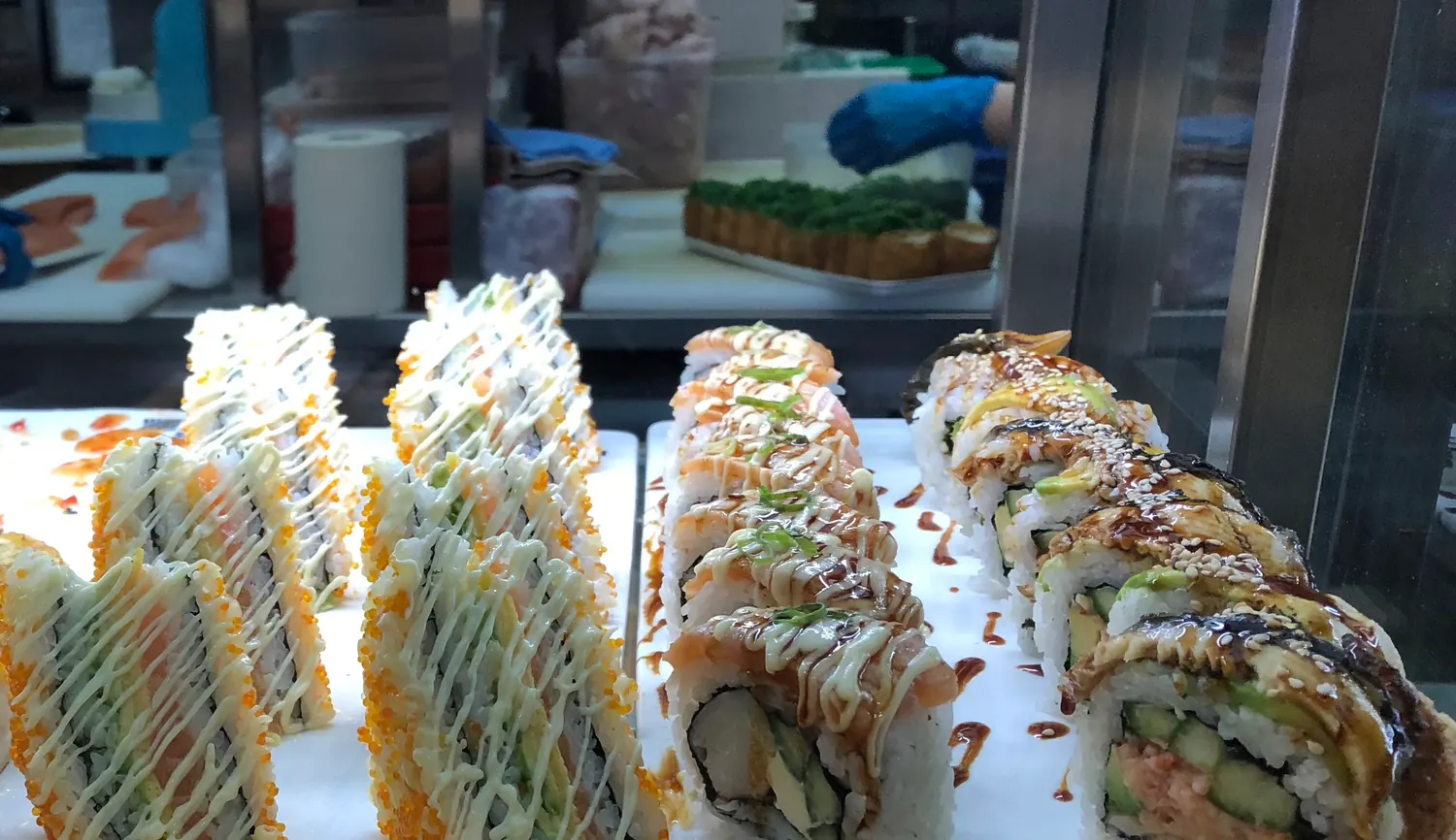 Rows of sushi rolls on trays behind a glass counter, sauce across the tops of them, with the kitchen working beyond the glass.