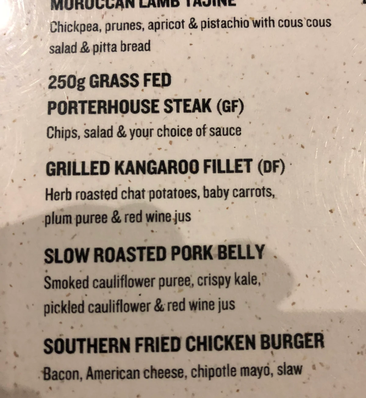 A printed menu board read close up: Moroccan lamb tagine, porterhouse steak, grilled kangaroo fillet, slow roasted pork belly and a fried chicken burger, each with a line of description under it.