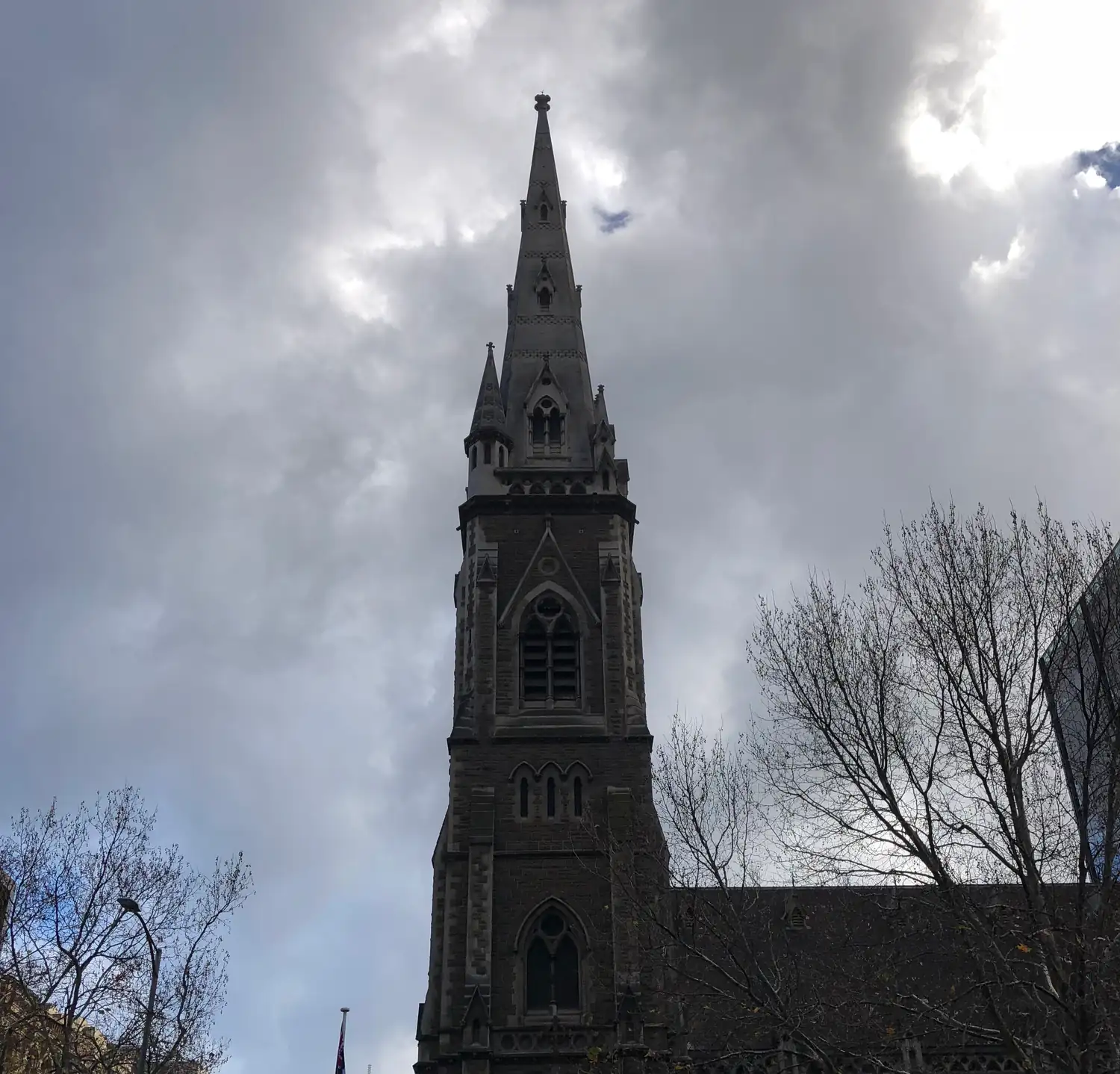 A church spire and tower in dark stone under heavy grey cloud, bare winter trees beside it.