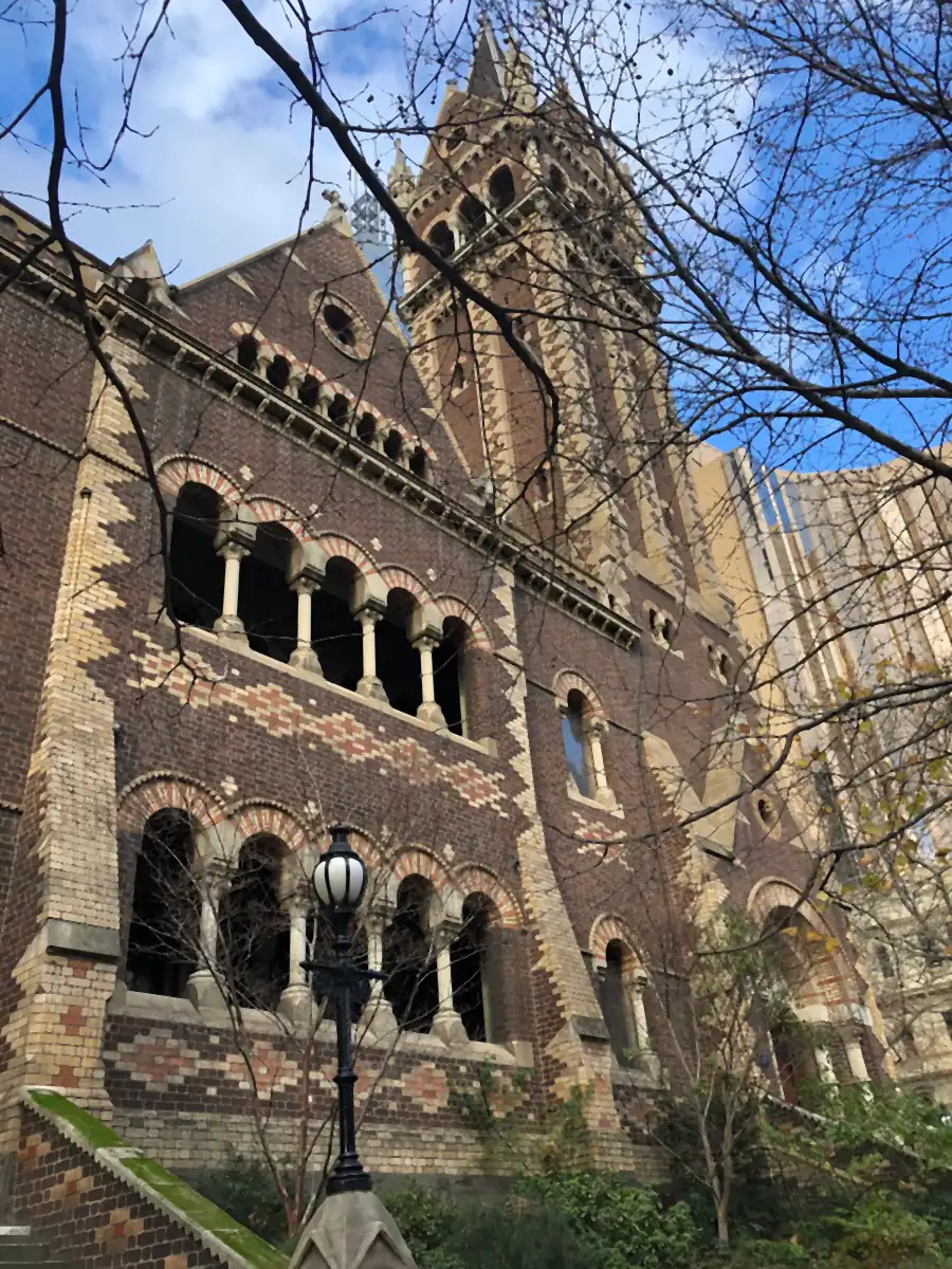 A Gothic revival building in polychrome brick, its tower and arched windows seen through bare winter branches.