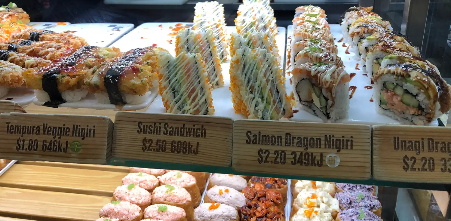 Trays of sushi and small cakes behind the glass of a food counter, with wooden dish cards in front.