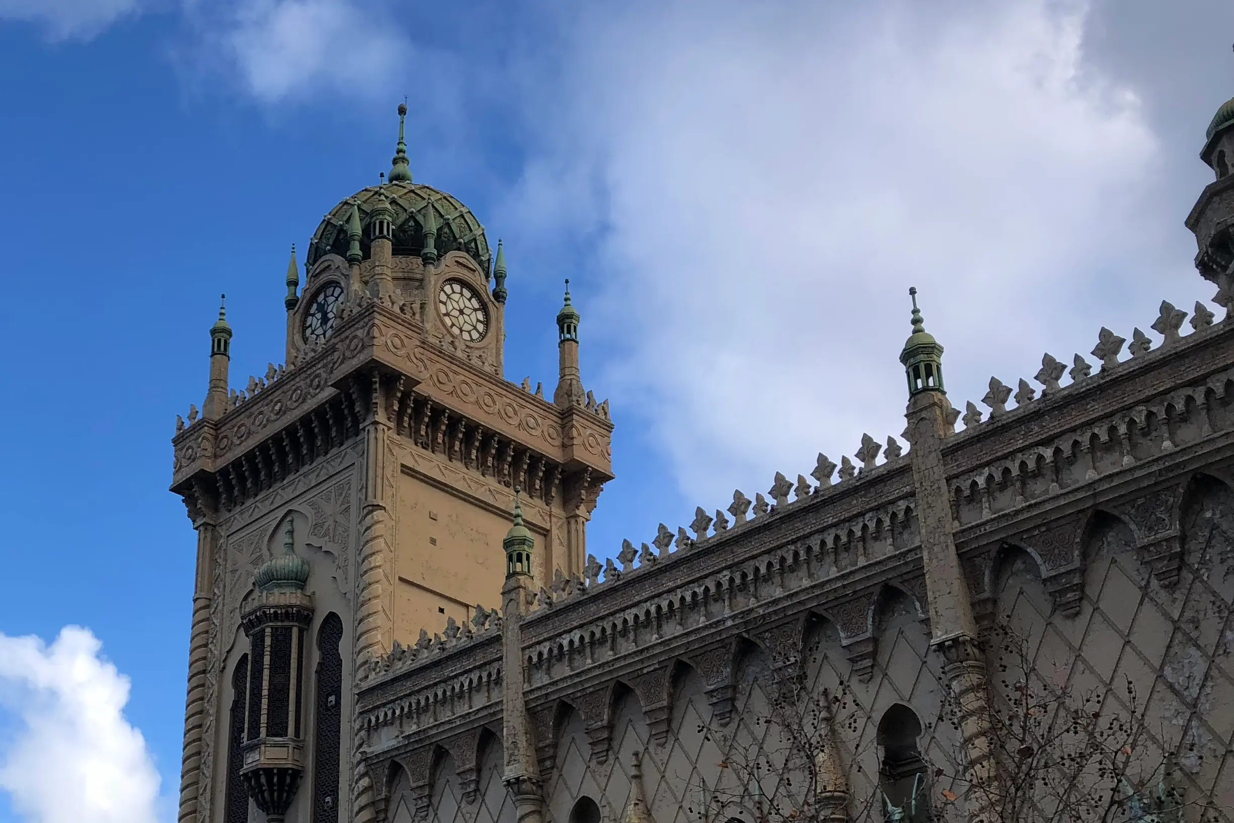 A green-domed clock tower above a stone facade patterned in arabesque arches, bare winter branches beside them against a deep blue sky.