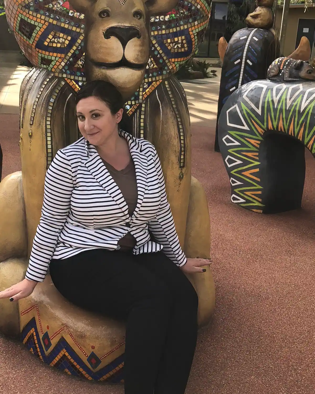 A woman in a striped jacket seated on a carved lion figure, mosaic covered animal sculptures standing around her in winter sun.