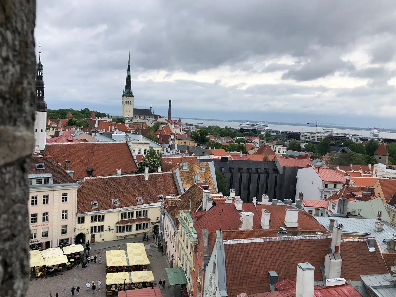 Red tiled roofs and pale houses seen from a tower, with a market square and its stalls below and a slender tower rising beyond the roofs.