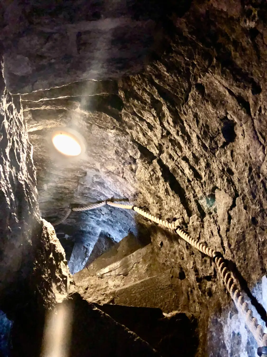 A passage cut through stone and lit from above, with a hand rail and a ladder.